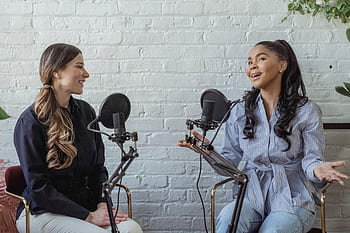 Two women engaged in a podcast recording session indoors, smiling and talking. HD wallpaper