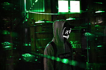 Man in a hoodie with glowing mask amidst neon green lights, evoking mystery and intrigue. HD wallpaper