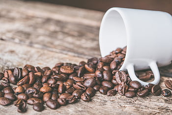 Coffee beans spilling from a white mug on a rustic wooden surface, embodying a cozy and warm atmosphere. HD wallpaper