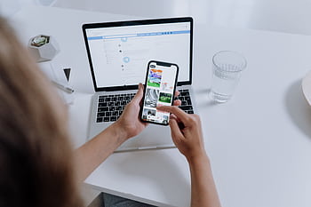 Person using both a smart and laptop, multitasking at a modern desk with office plants. HD wallpaper