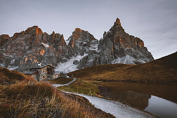 Tranquil mountain cabin scene under dramatic sunset light, featuring rugged peaks and serene lake reflection. HD wallpaper