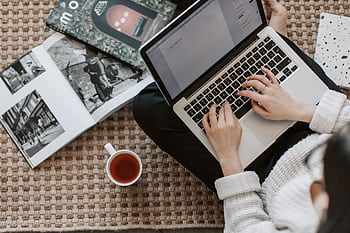 Overhead view of a woman typing on a laptop at home, surrounded by magazines and a cup of tea on the floor. HD wallpaper