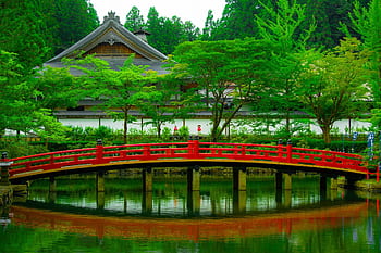 Serene Japanese garden with traditional architecture and a vibrant red bridge over a reflective pond. HD wallpaper