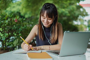 Smiling young woman studying at outdoor table with a laptop and notebook, listening to music with ears. HD wallpaper