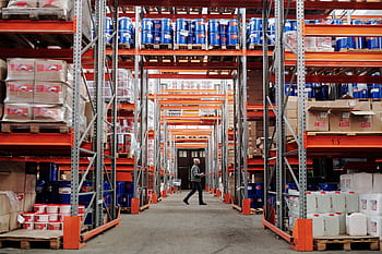 A man walking through a large industrial warehouse with stacked shelves filled with goods and products. HD wallpaper
