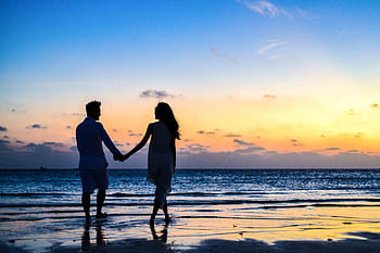 A romantic couple walks hand in hand on a tropical beach at sunset, enjoying a serene moment together. HD wallpaper