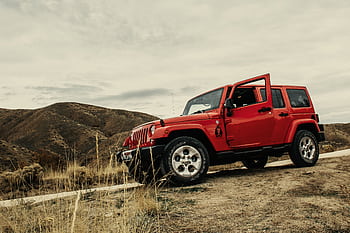 A vibrant red SUV parked on a dirt road with a mountain landscape backdrop under a cloudy sky. HD wallpaper