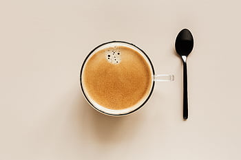 A minimalist shot of a cup of coffee with foam next to a spoon on a beige backdrop. HD wallpaper