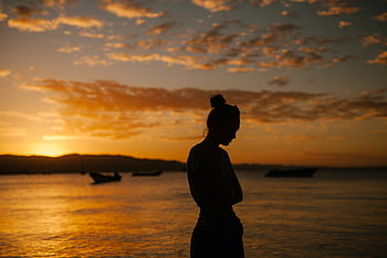 A silhouette of a woman standing by the sea during a serene sunset, exuding tranquility and solitude. HD wallpaper