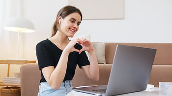 A young woman smiling during a video call, showing a heart gesture with her hands in a cozy living room. HD wallpaper