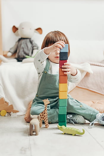A cute child stacks colorful wooden blocks while playing indoors, surrounded by toys. HD phone wallpaper