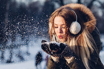 A young woman enjoying a snowy day, blowing snow while wearing cozy earmuffs and a winter coat. HD wallpaper