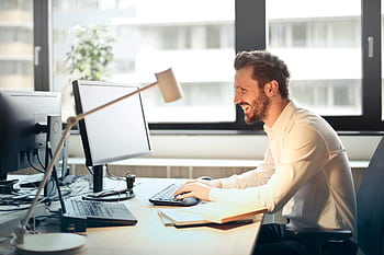 A man smiling while working at an office desk with a computer and natural daylight streaming in through large windows. HD wallpaper
