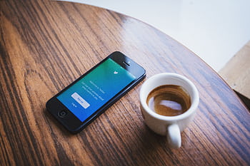 Close-up of a smart with the Twitter app next to a cup of coffee on a wooden table. HD wallpaper