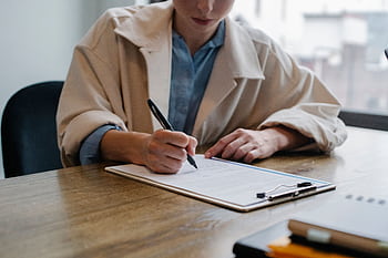 A woman in a formal setting fills out paperwork on a clipboard at an office desk. HD wallpaper