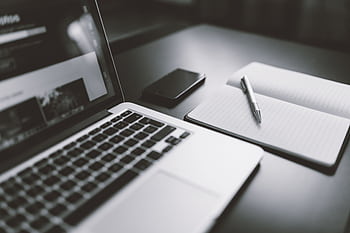 A sleek black and white workspace featuring a laptop, smart, and notebook with a pen, highlighting modern technology. HD wallpaper