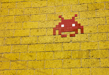 Street art featuring a retro pixelated space invader on a vivid yellow brick wall. HD wallpaper