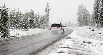 A car drives on a snowy road surrounded by trees during a snowstorm, creating a wintry scene. HD wallpaper