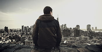 Man sitting on rooftop overlooking city skyline, contemplating the urban landscape. HD wallpaper