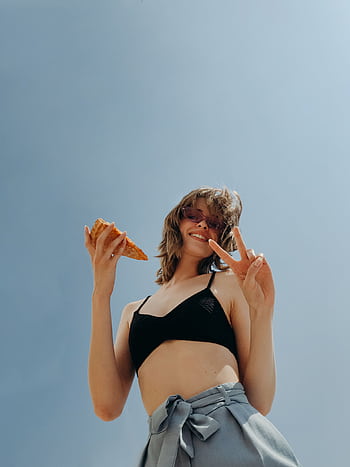Smiling woman in swimwear posing outdoors with peace sign and slice of pizza under bright summer sky. HD phone wallpaper