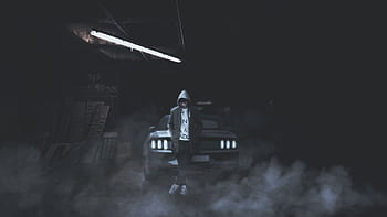 A hooded person stands confidently against a car in a foggy, dimly lit garage. HD wallpaper