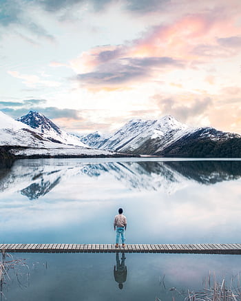 Person standing by a serene lake with snow-capped mountains in Otago, New Zealand. HD phone wallpaper