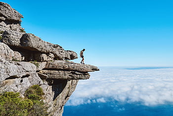 A lone person on a dramatic cliff edge gazing over a sea of clouds under a bright blue sky. HD wallpaper