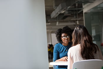 Two women engaged in a discussion in a modern office environment, highlighting communication and interaction. HD wallpaper