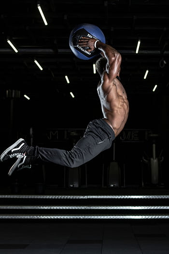 Muscular athlete performing a high jump with a medicine ball in a gym, showcasing strength and fitness. HD phone wallpaper