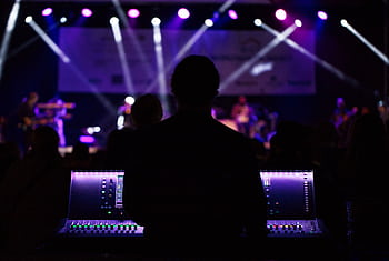 A sound engineer manages the audio mixer during a vibrant live concert with colorful lighting. HD wallpaper