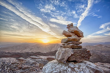 Stacked stones atop a mountain at sunset, with a breathtaking sky and landscape in the background. HD wallpaper