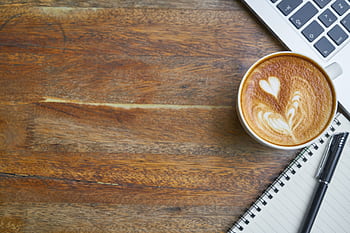Cozy workspace featuring a cappuccino with latte art, open notebook, and laptop on a wooden desk. HD wallpaper