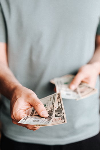 Close-up of an unrecognizable man holding dollar bills with a blurred background indoors. HD phone wallpaper