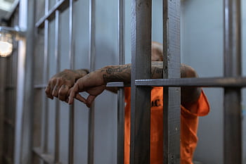 A tattooed inmate's arms rest on jail cell bars, wearing an orange uniform inside a prison. HD wallpaper
