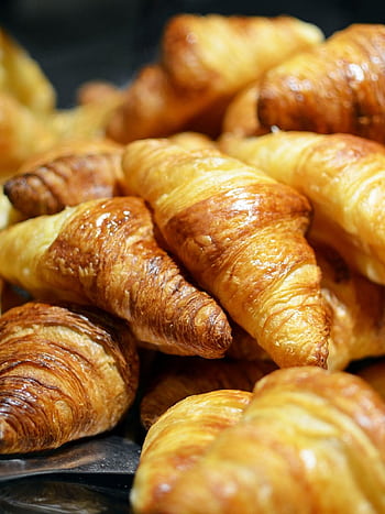 A stack of golden, freshly baked croissants showcasing flaky layers in a bakery setting. HD phone wallpaper