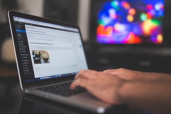 Hands typing on a laptop with a blog post visible, cozy indoor setting with colorful screen in background. HD wallpaper