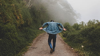 A man in denim jacket hikes along a foggy trail surrounded by lush greenery, creating a sense of adventure. HD wallpaper