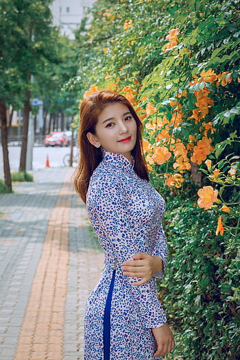 Asian woman in a floral Ao Dai posing beside vibrant orange flowers outdoors. HD phone wallpaper