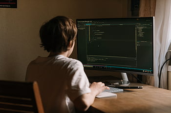 A teenager focused on coding software on a monitor in a home office setting. HD wallpaper