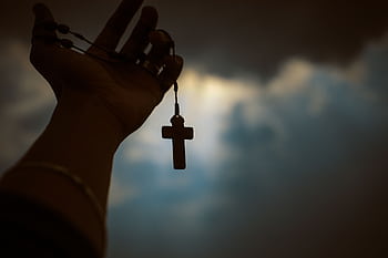 Silhouette of a hand holding a rosary with a cross against a dramatic sky, symbolizing faith and spirituality. HD wallpaper