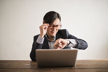 Asian businessman checking watch while working on laptop, expressing stress and urgency. HD wallpaper