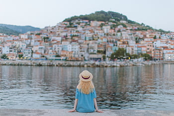 A woman in a blue dress sits on a pier, gazing at a que coastal town. HD wallpaper