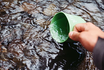 Hand collecting fresh water from a clear stream using a green cup in a natural setting. HD wallpaper