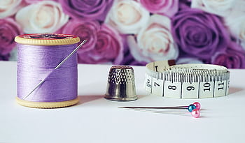 A close-up of sewing tools including lilac thread, a thimble, tape measure, and colorful pins with a rose backdrop. HD wallpaper