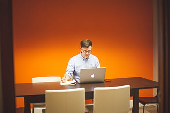 Young man working on a laptop in a stylish office space with an orange wall. HD wallpaper