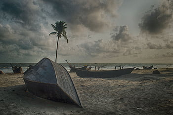 Tranquil beach scene with wooden boats, palm tree, and cloudy sky in Cox's Bazar. HD wallpaper