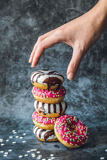 A hand reaches for a stack of colorful donuts with chocolate drizzle and sprinkles, tempting and delicious. HD phone wallpaper