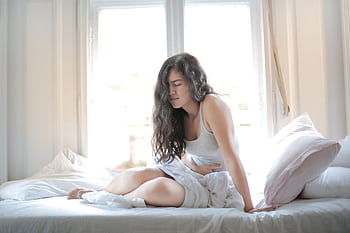 Woman sitting on bed holding her stomach, showing signs of discomfort or pain indoors. HD wallpaper