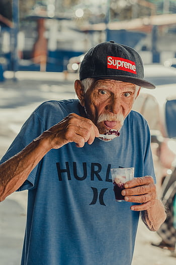 Senior man savoring a sweet treat outdoors, wearing a trendy cap and casual outfit. HD phone wallpaper
