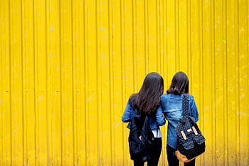 Two women wearing denim jackets and backpacks stand against a vibrant yellow wall. HD wallpaper